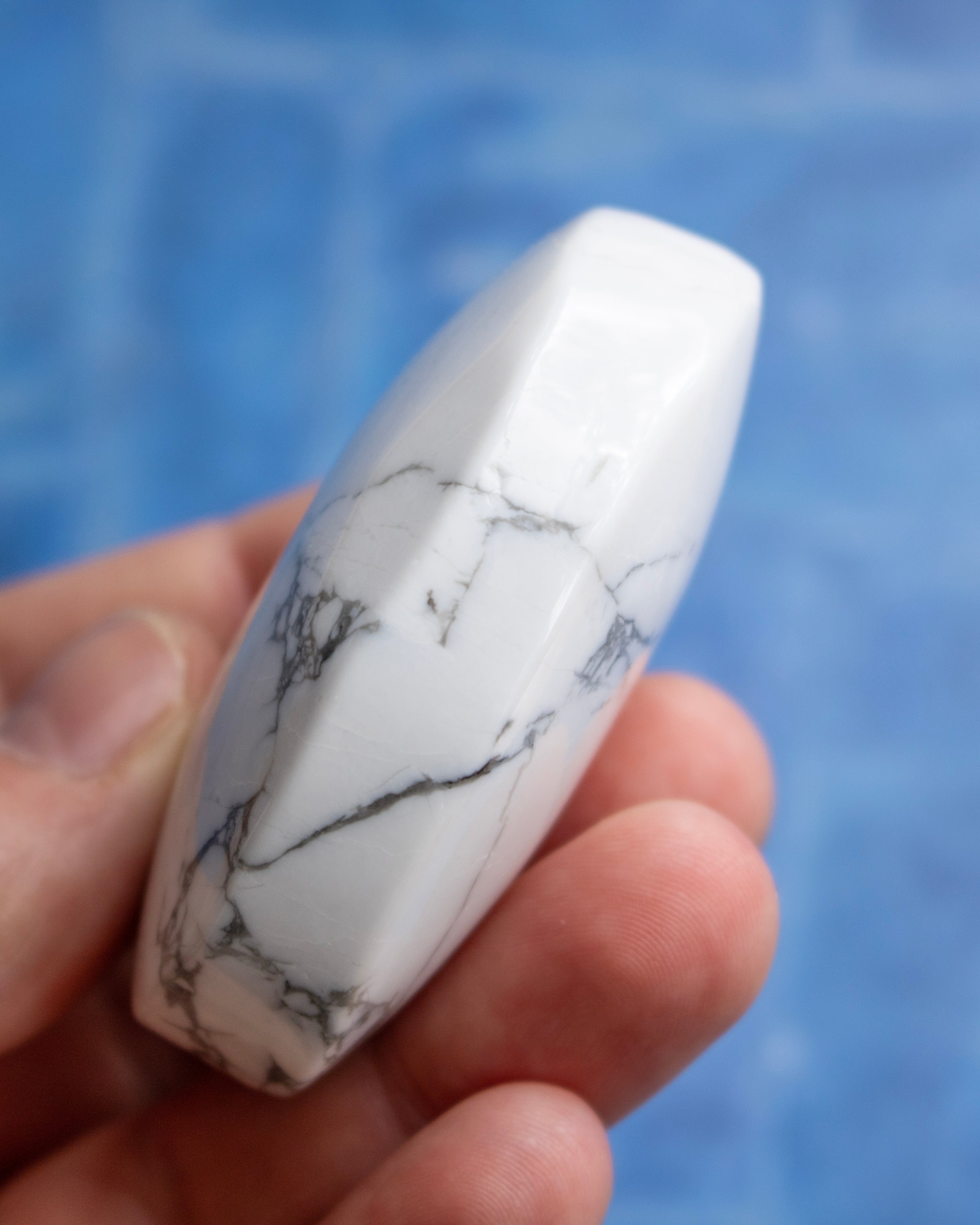 A hand holding a polished white agate bead with natural gray and black veining, set against a blurred blue background.