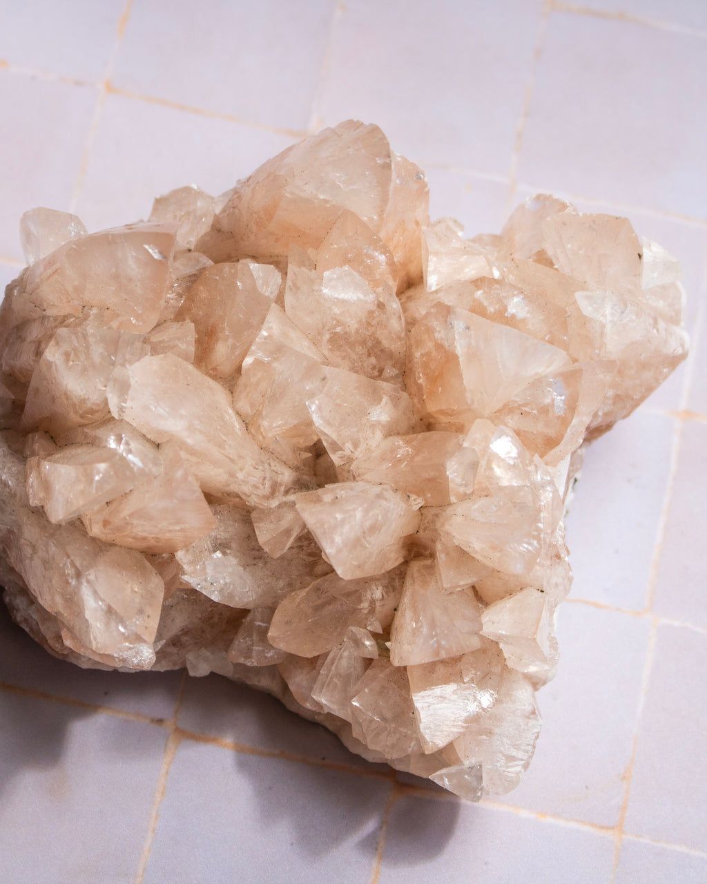 A raw crystal cluster with natural rough edges and facets, showing pale peach and translucent white tones, resting on a light tiled surface.