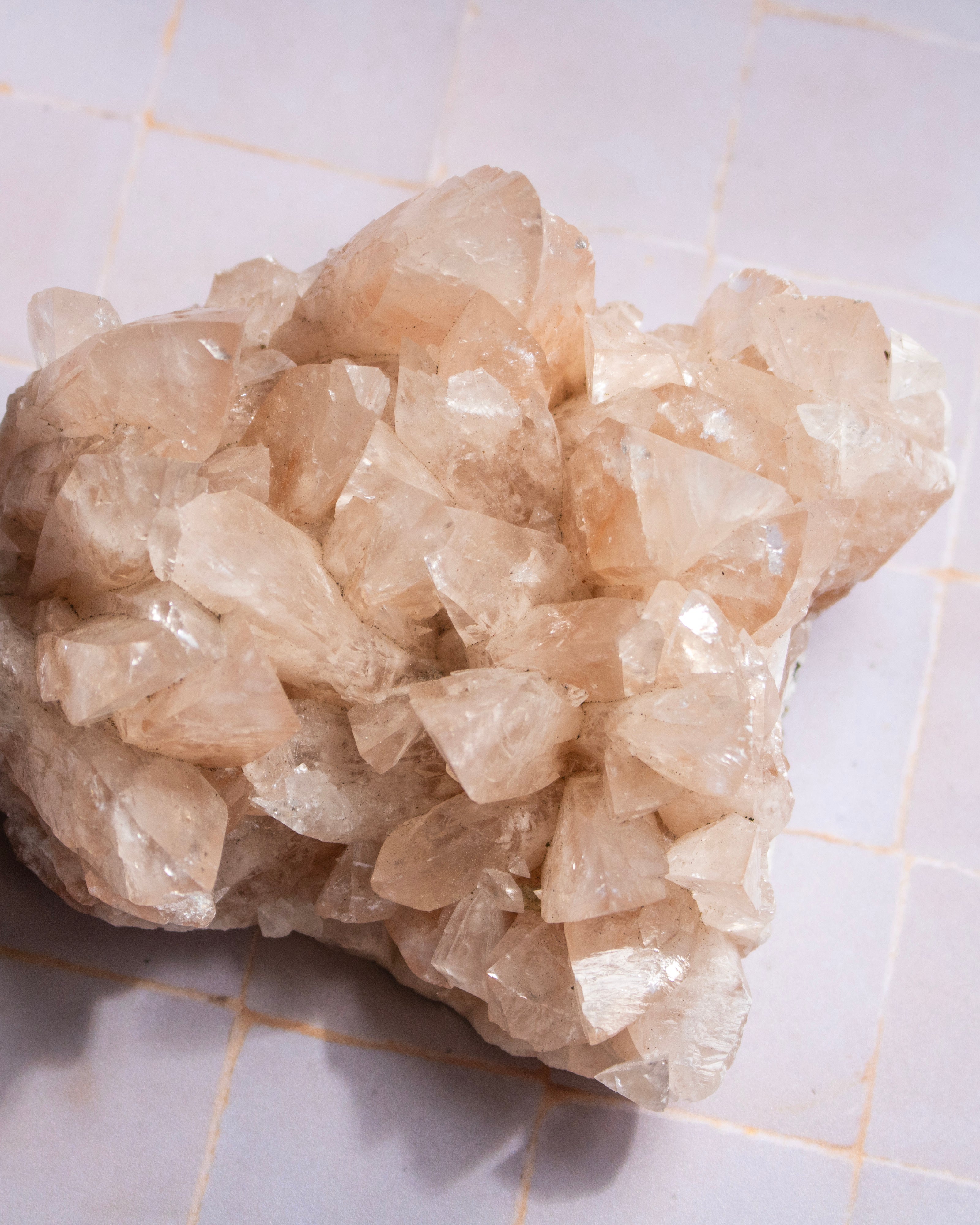 A raw crystal cluster with natural rough edges and facets, showing pale peach and translucent white tones, resting on a light tiled surface.