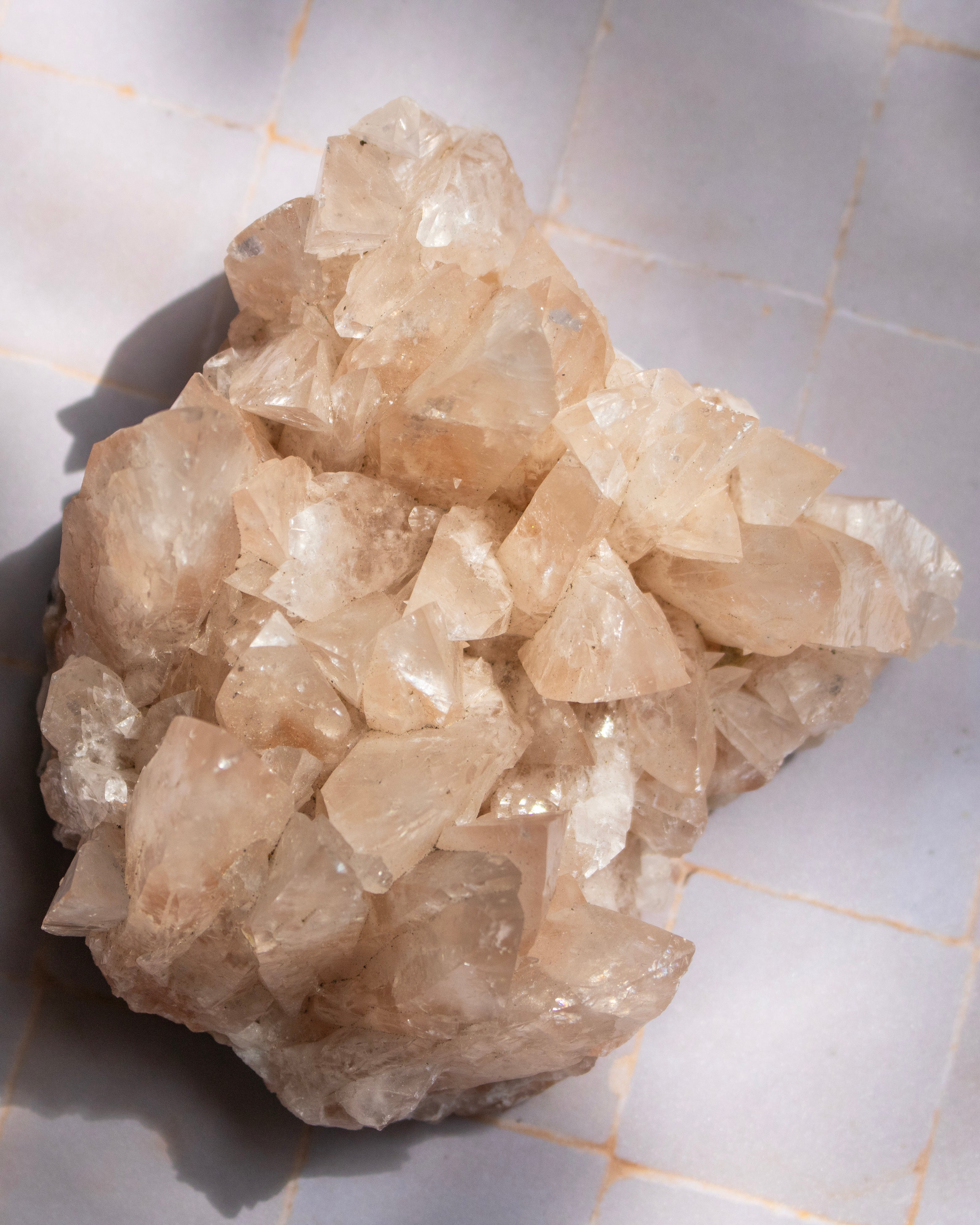 A raw crystal cluster with translucent pale peach and cream quartz-like formations, photographed on a light tiled surface with strong natural shadows.
