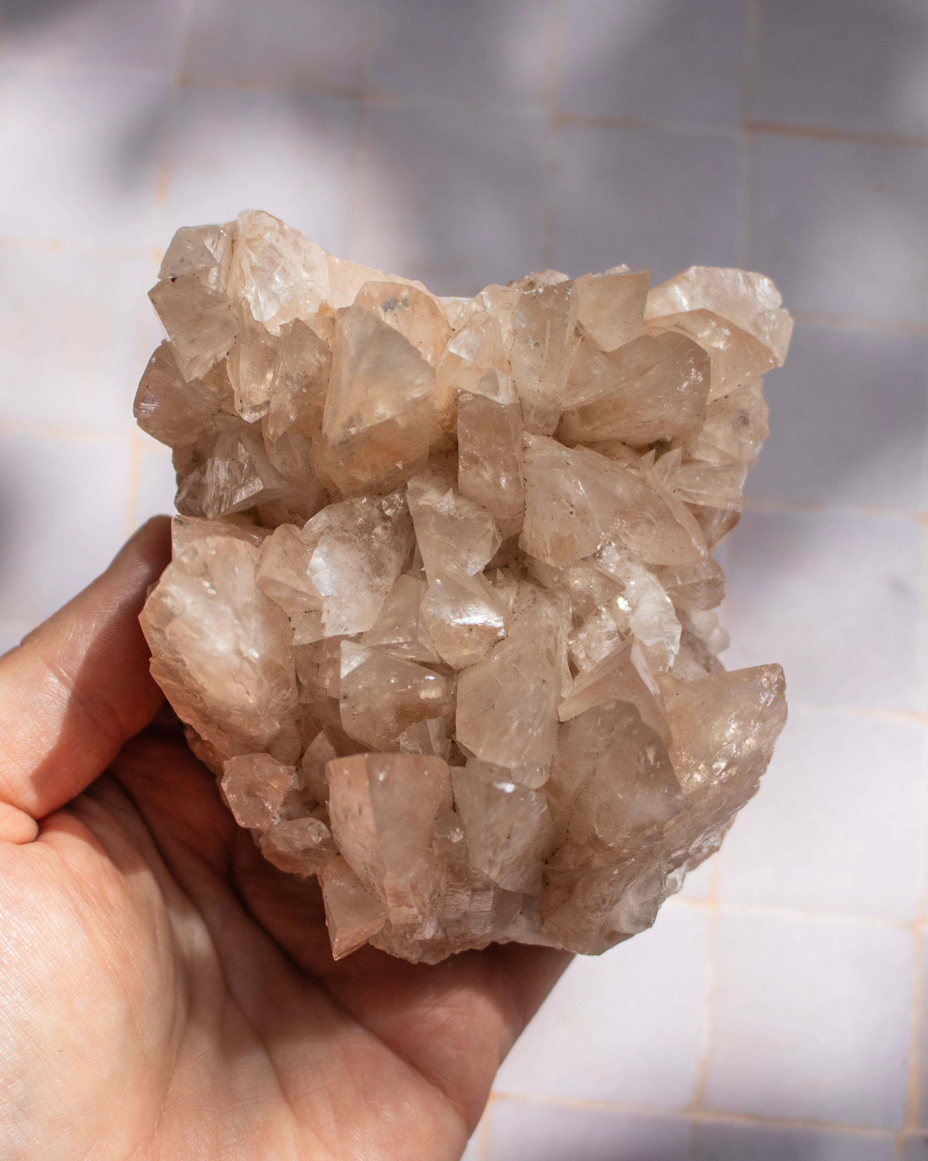 A hand holding a raw crystal cluster with natural rough edges and translucent pale pinkish-tan facets, set against a tiled wall with soft shadows.