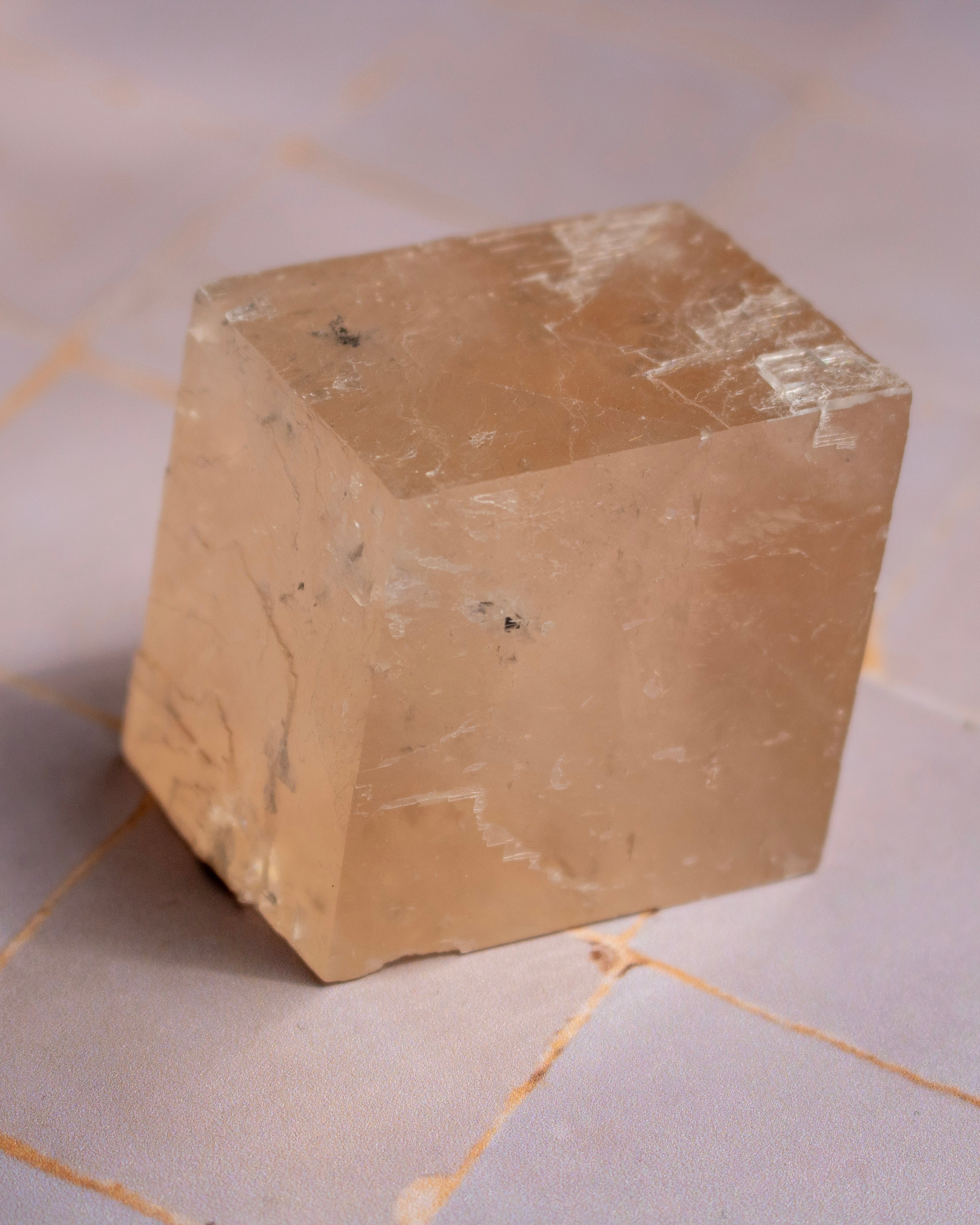 A translucent pale peach-beige crystal cube with natural striations and inclusions, photographed on a light stone-tiled surface.