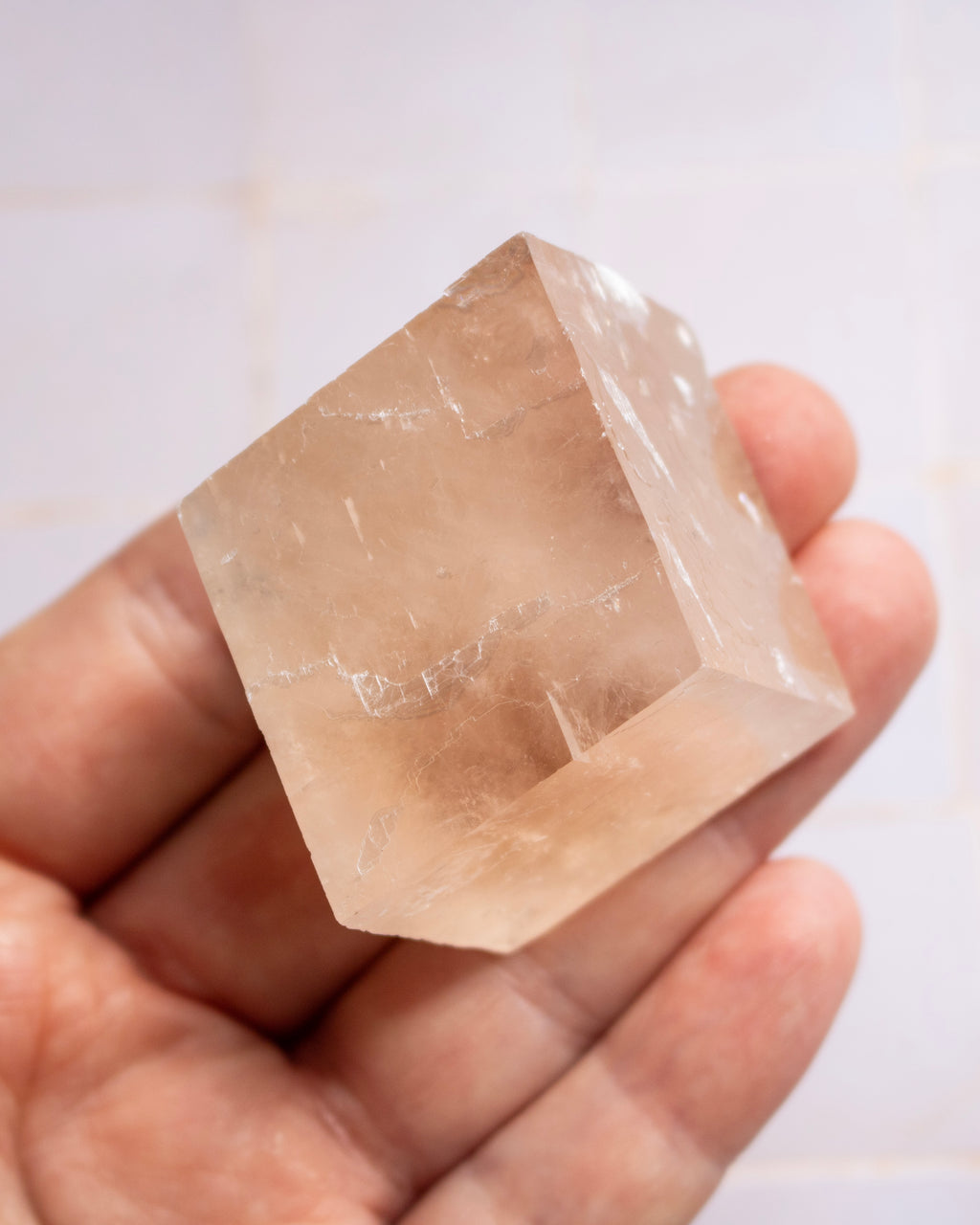 A hand holding a faceted, translucent pale peach crystal or quartz cube against a light tiled background.