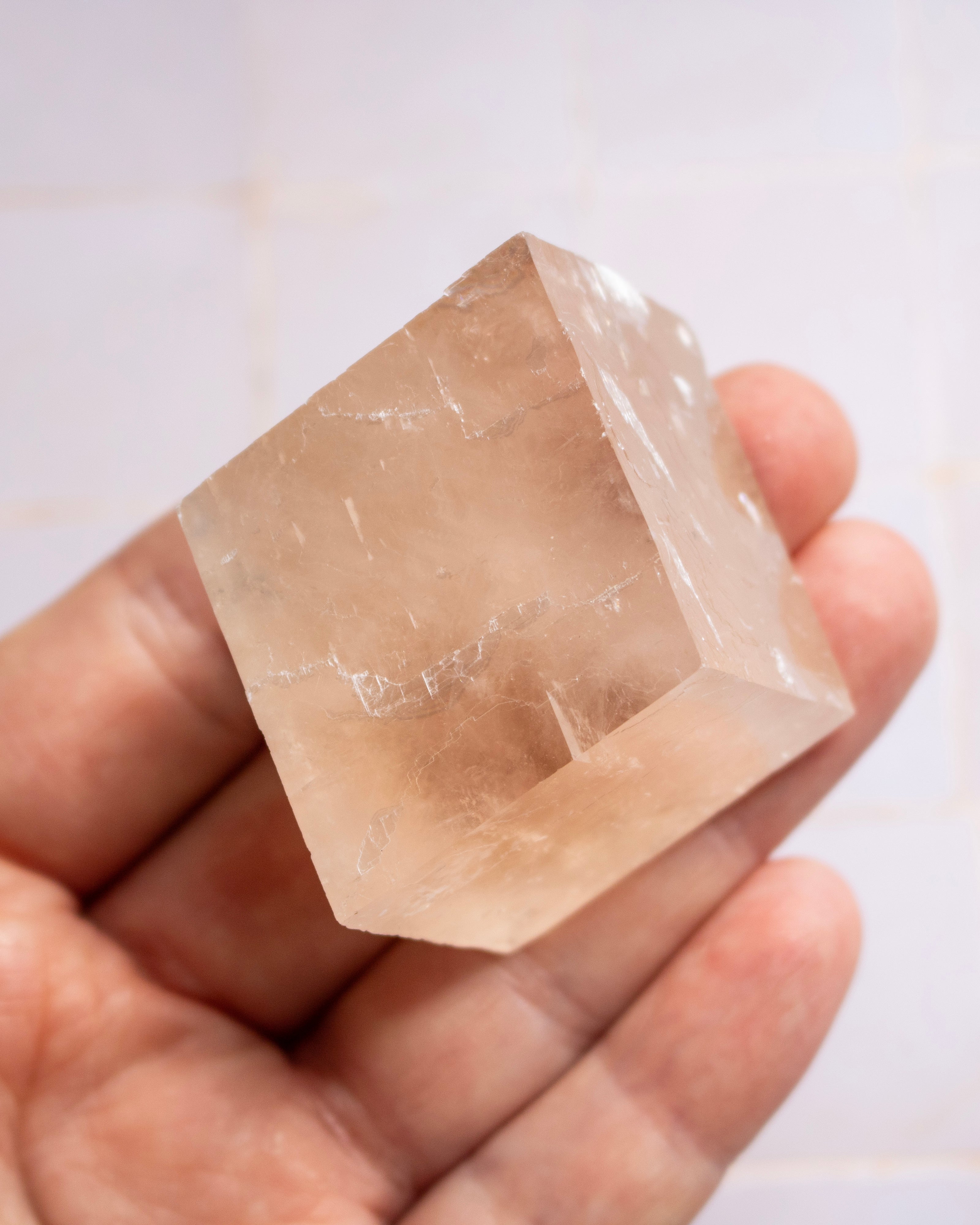 A hand holding a faceted, translucent pale peach crystal or quartz cube against a light tiled background.