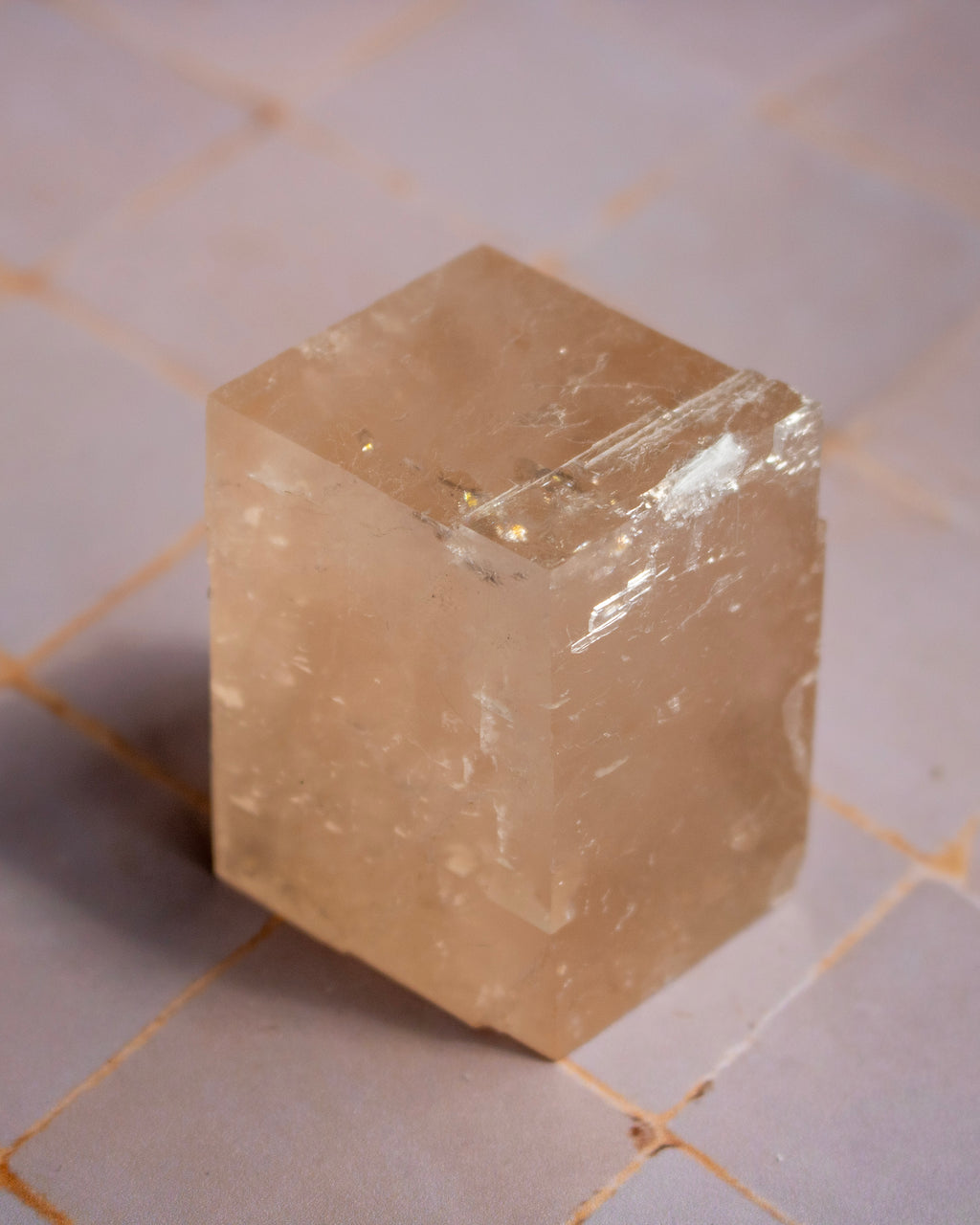 A translucent pale peach-colored crystal cube with natural internal inclusions and rough edges, standing on a light tiled surface.
