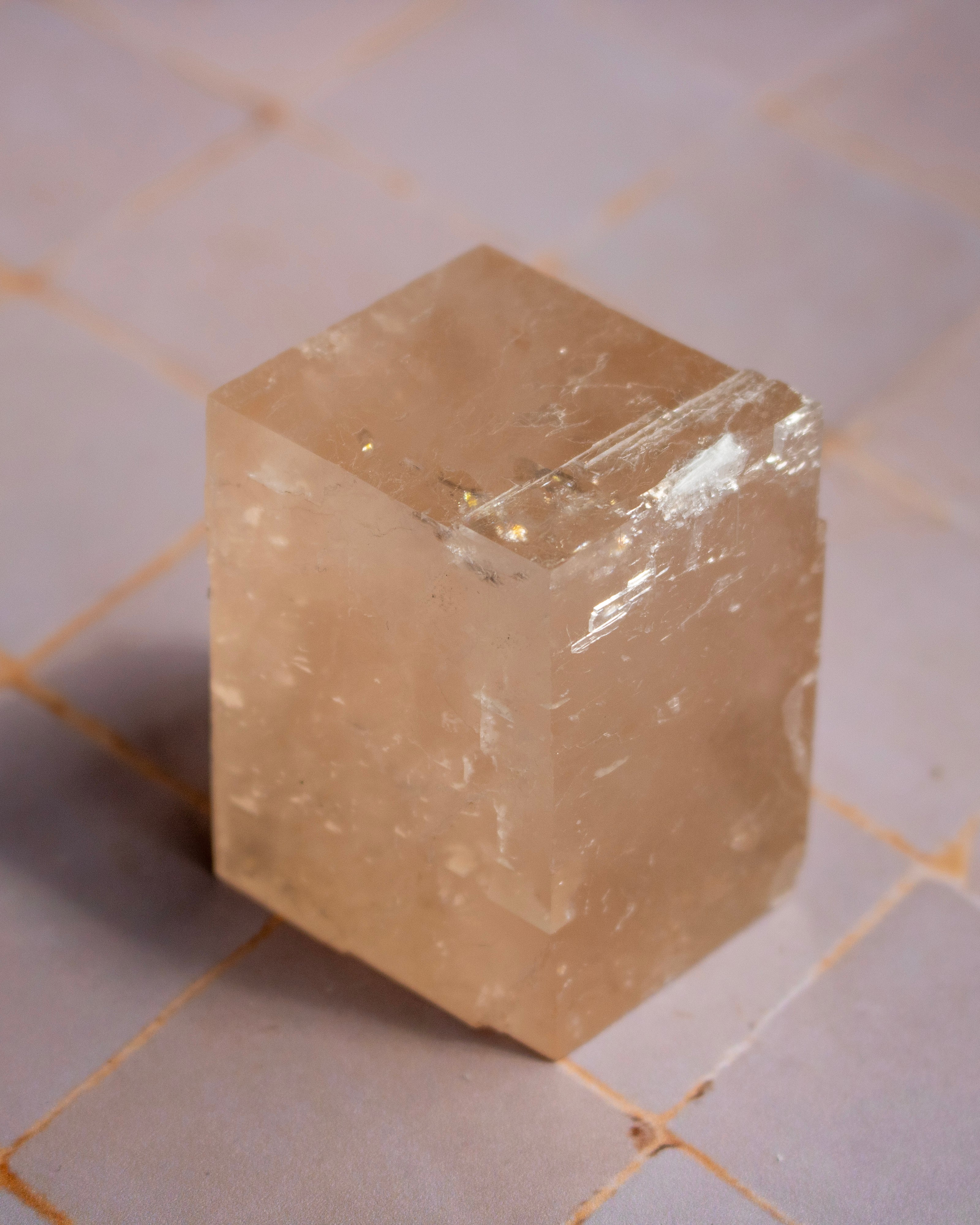 A translucent pale peach-colored crystal cube with natural internal inclusions and rough edges, standing on a light tiled surface.