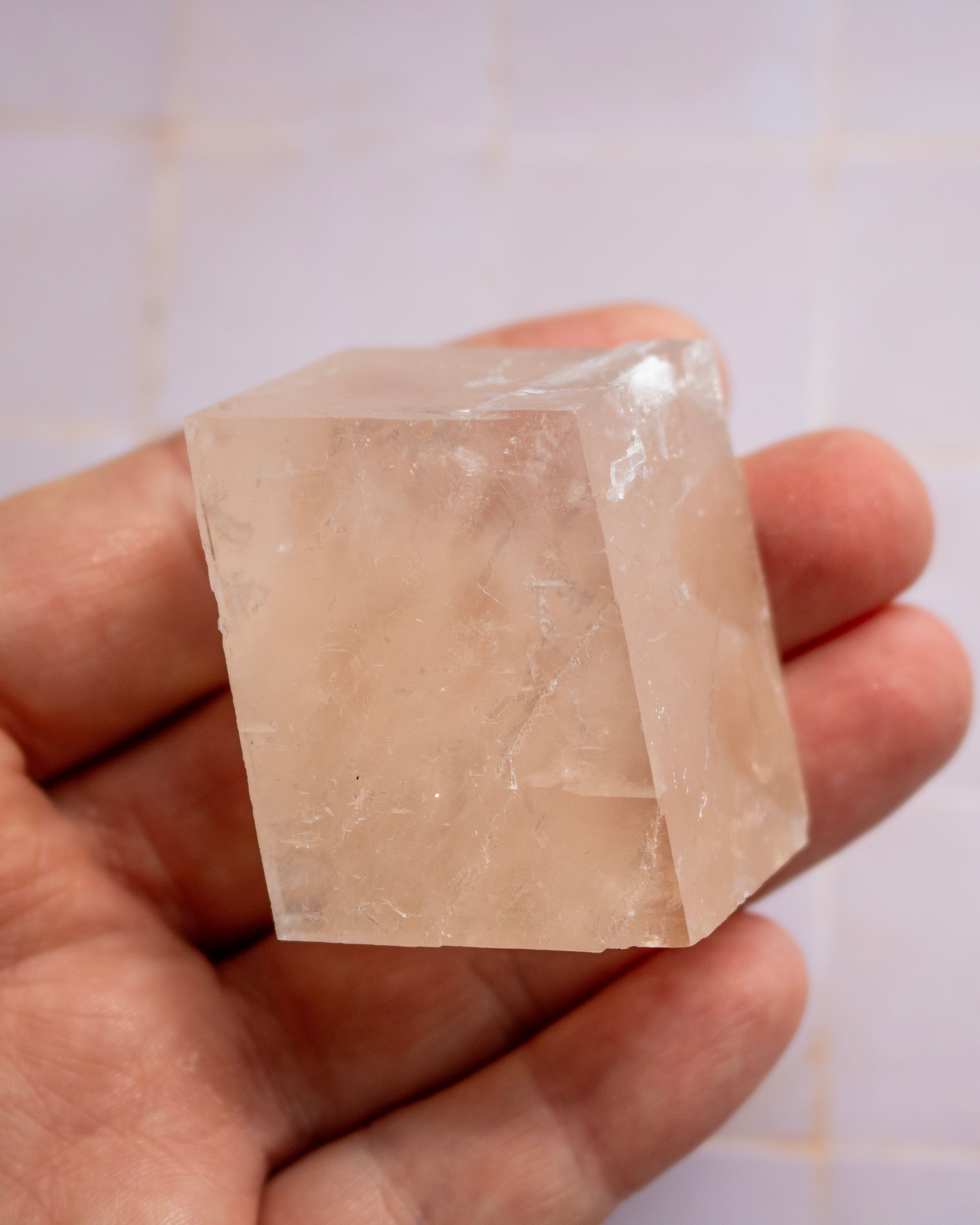 A hand holding a translucent pale pink crystal cube against a light tiled background.
