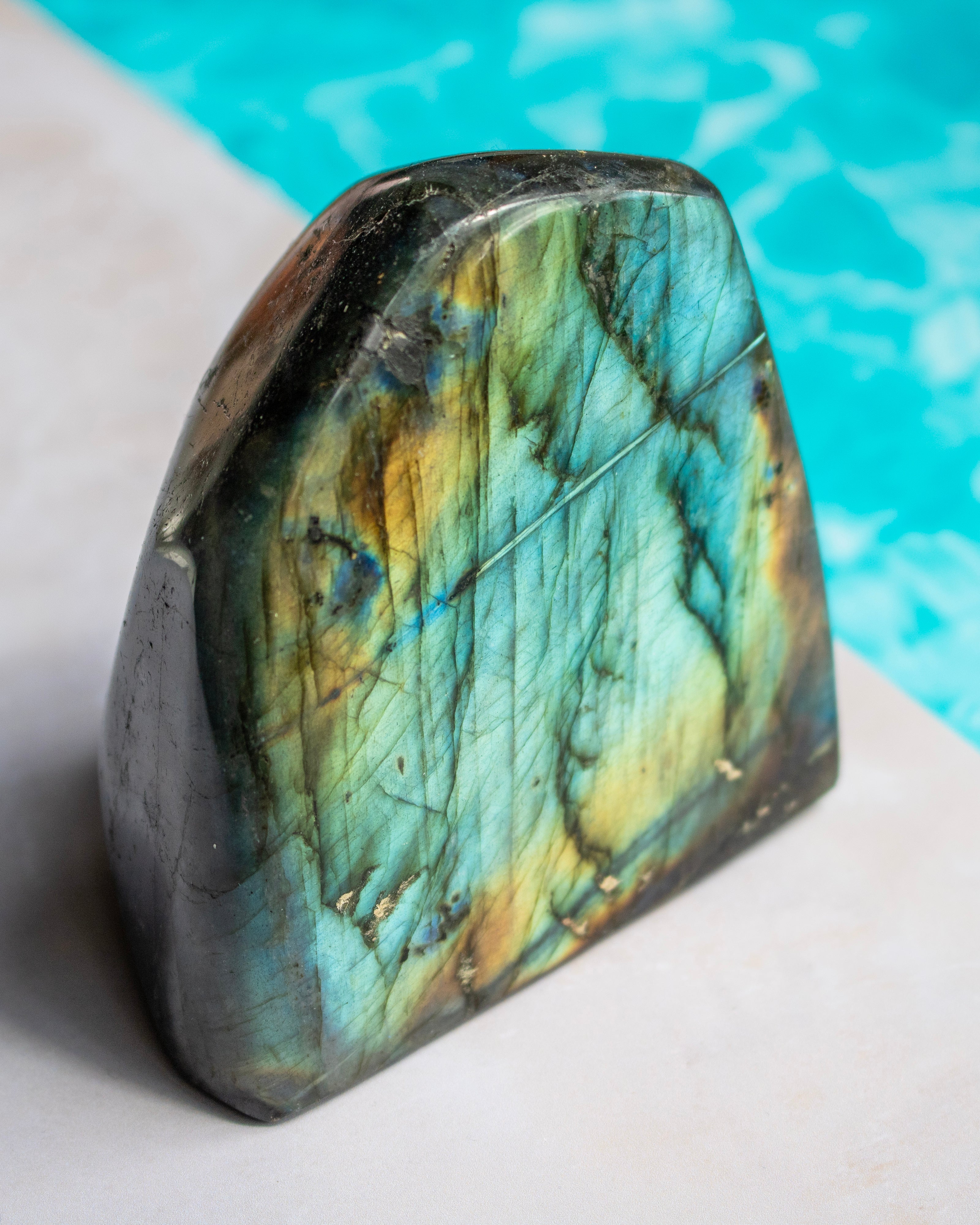 A polished labradorite stone with a natural irregular shape, displaying vibrant green, blue, and gold iridescence, resting on a light surface with a turquoise pool in the background.