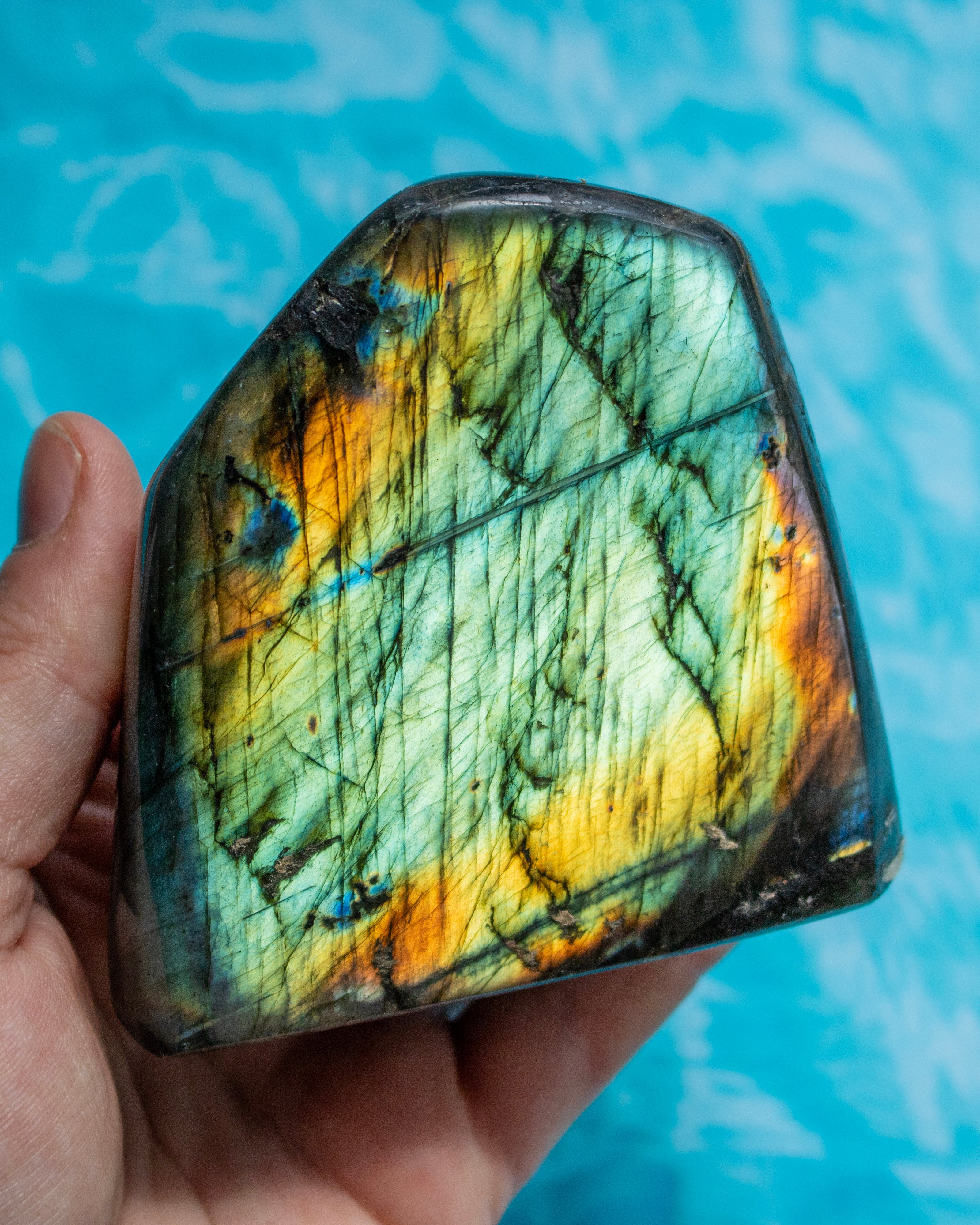 A hand holds a polished labradorite stone showing a vibrant play-of-color with green, yellow, orange, and dark gray, set against a bright blue water background.