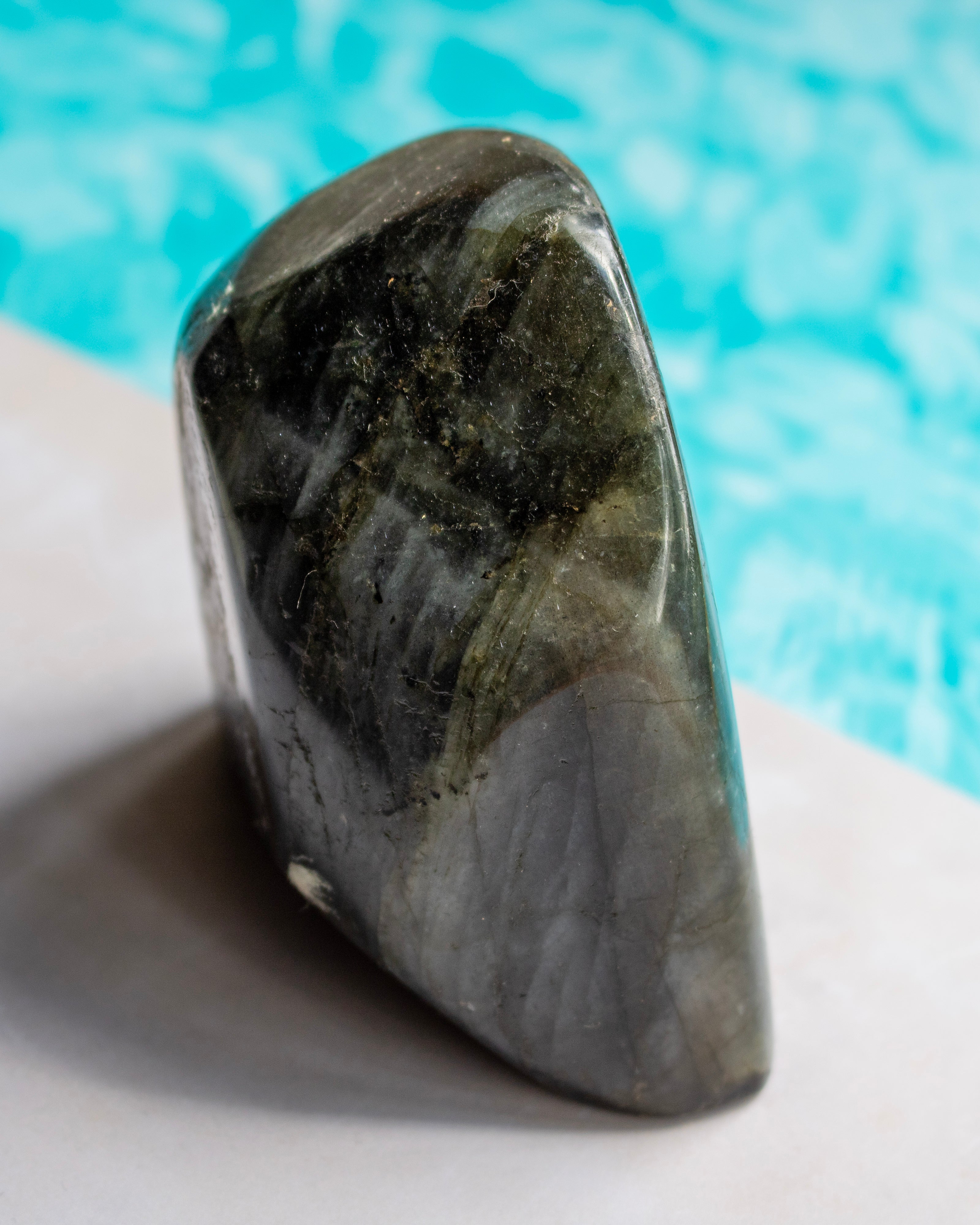 A polished natural stone with a dark gray and olive-green marbled pattern, standing upright on a light surface with a bright turquoise blue background.