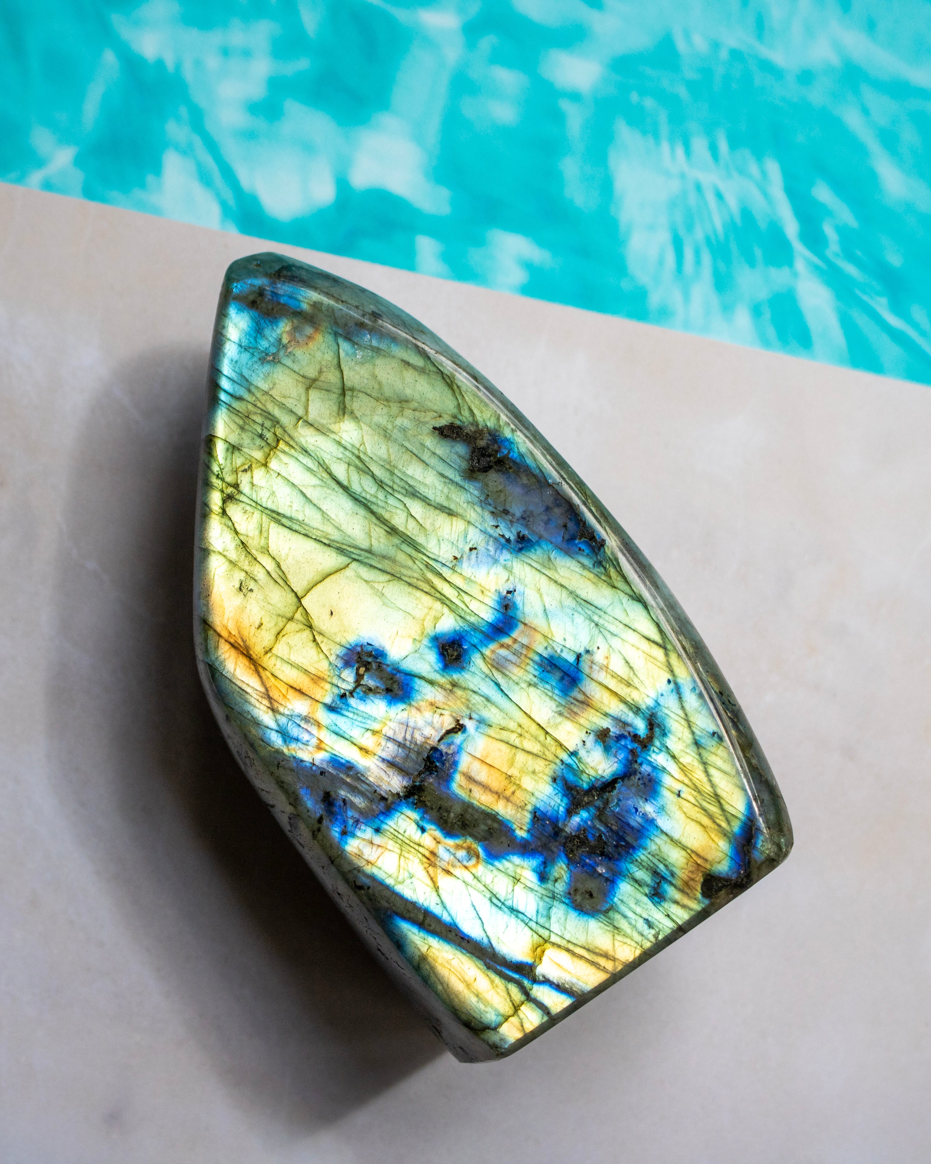 A polished labradorite stone with vibrant blue, gold, and green iridescent flashes, resting on a light beige surface with a turquoise pool background.