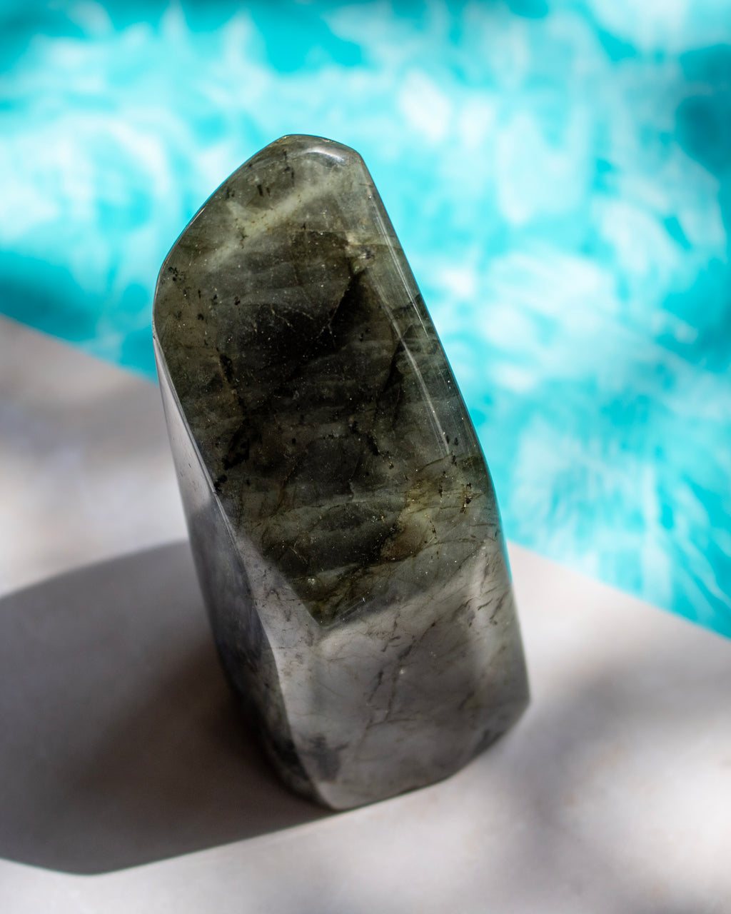 A polished natural crystal or stone tower with a pointed top, showing dark gray and olive-green internal banding, standing on a light surface with a bright turquoise blurred background.
