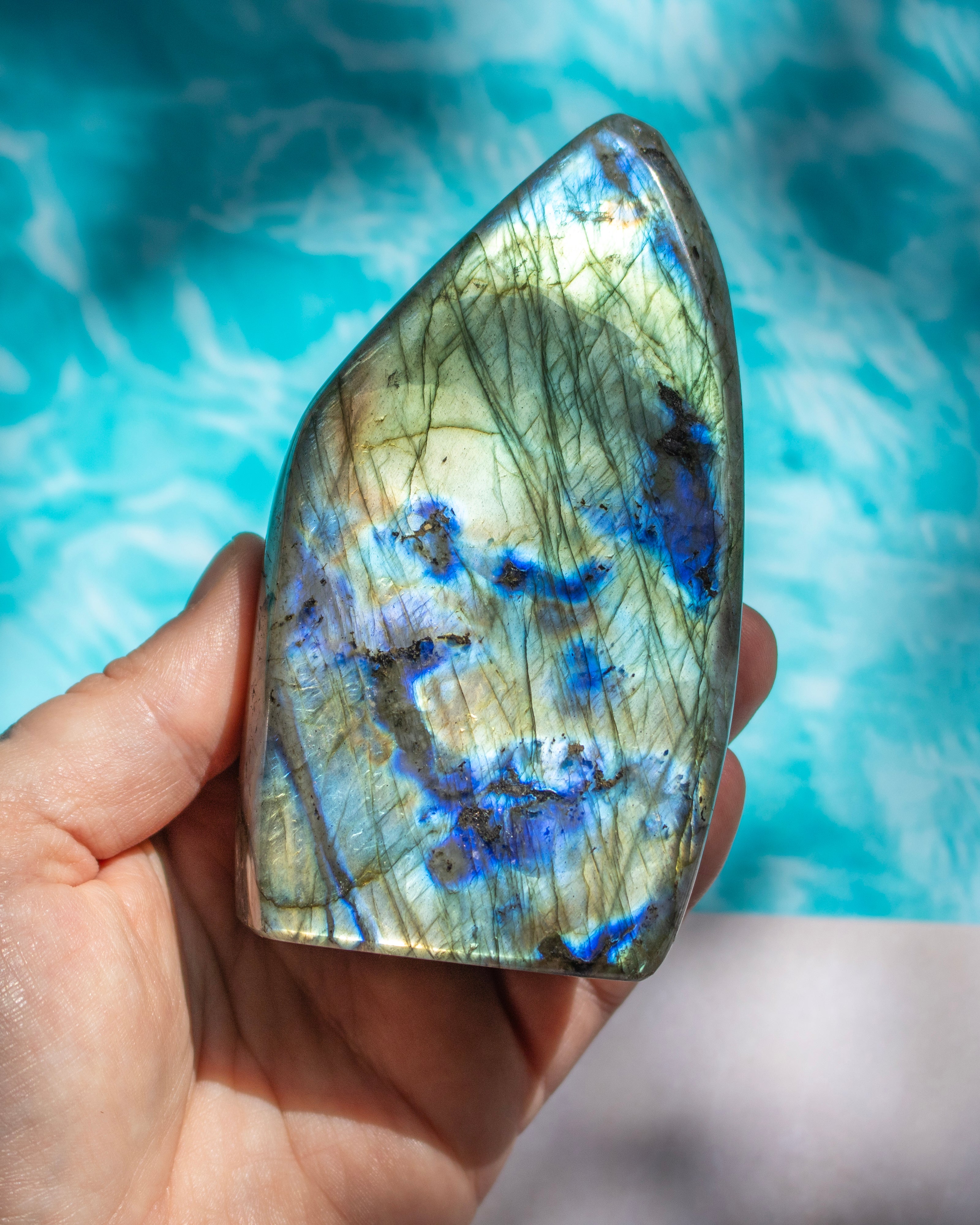 A hand holding a polished labradorite crystal stone with vibrant blue flashes and golden-green needle-like inclusions, set against a blurred turquoise background.