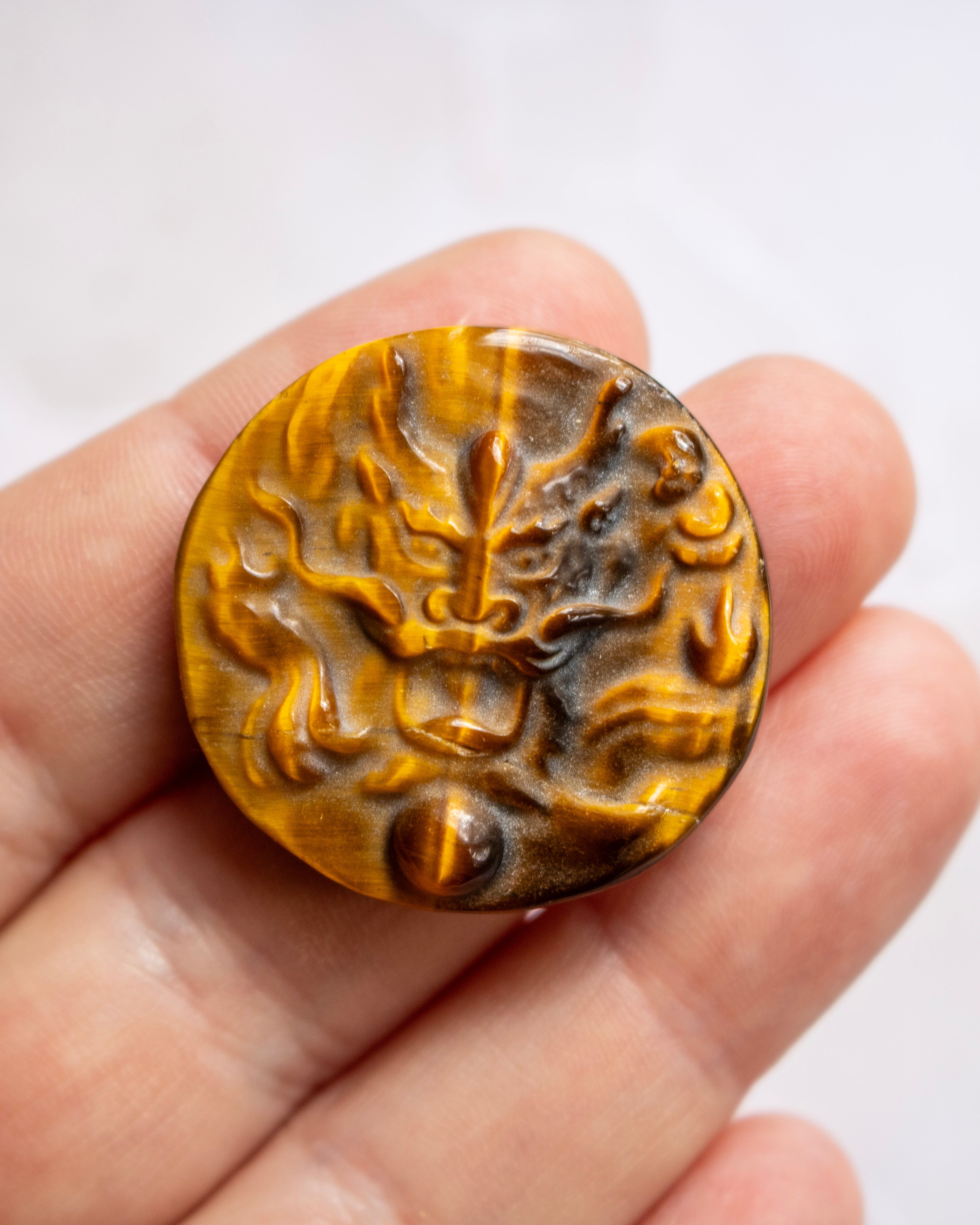 A close-up of a hand holding a round, carved amber-brown button with an embossed dragon design, set against a plain light background.