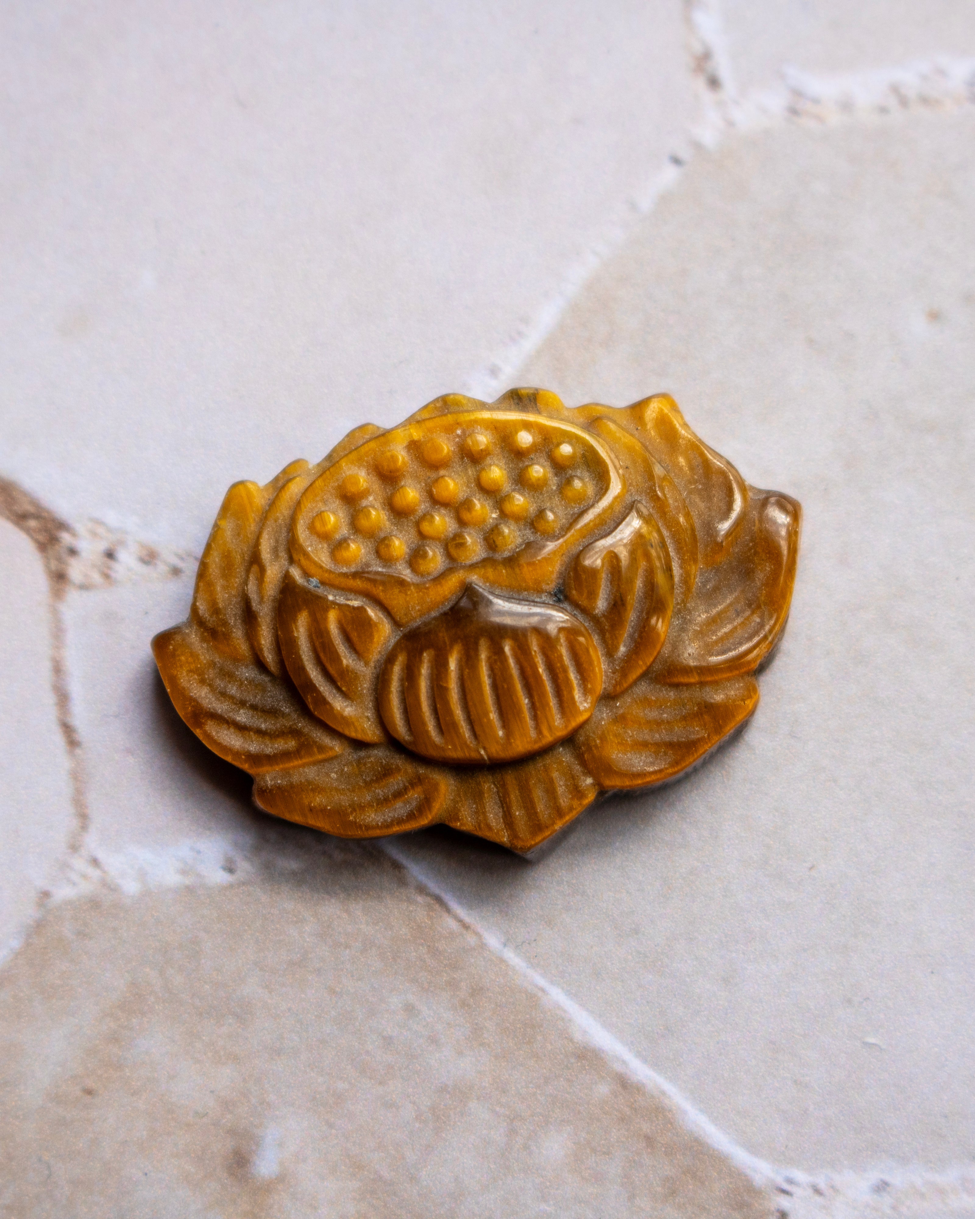 A carved brown lotus flower pendant or ornament with detailed ridges and a dotted center, resting on a light beige stone surface.