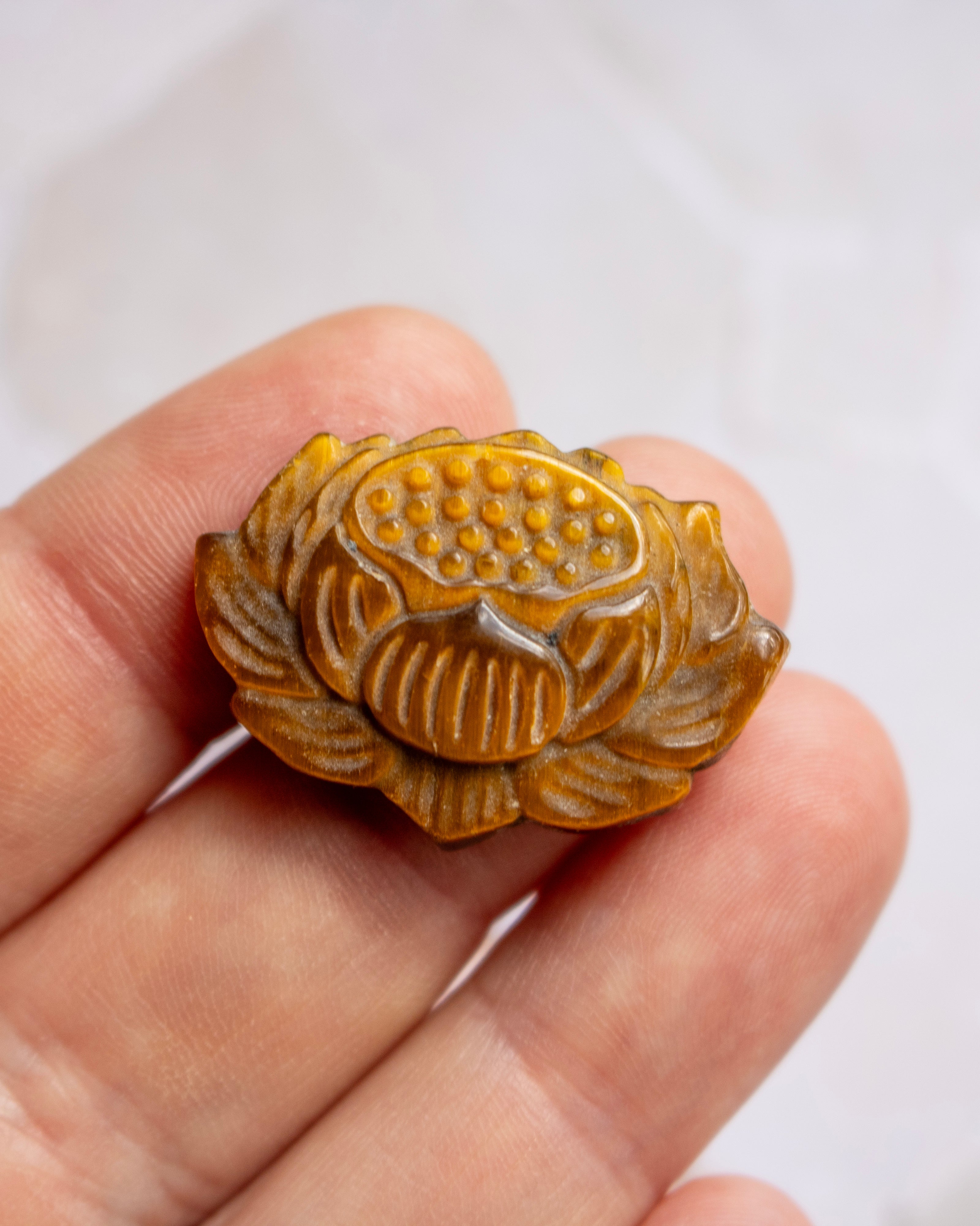A close-up of a hand holding a small carved lotus-shaped pendant or charm in warm amber-brown tones against a soft light gray background.