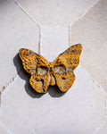 A butterfly-shaped pendant with a textured, organic pattern in golden yellow and brown, resting on a light beige stone surface.
