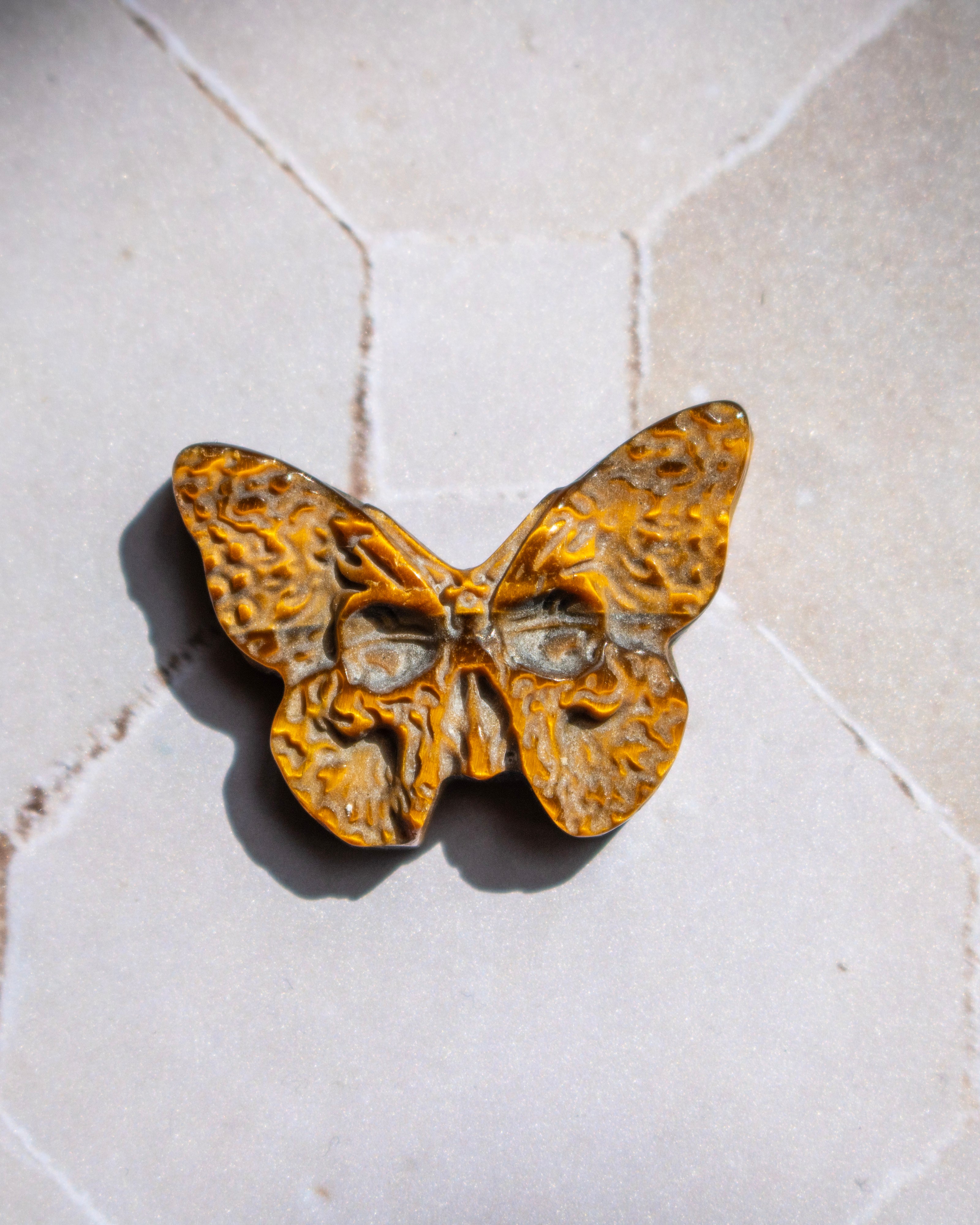 A butterfly-shaped pendant with a textured, organic pattern in golden yellow and brown, resting on a light beige stone surface.