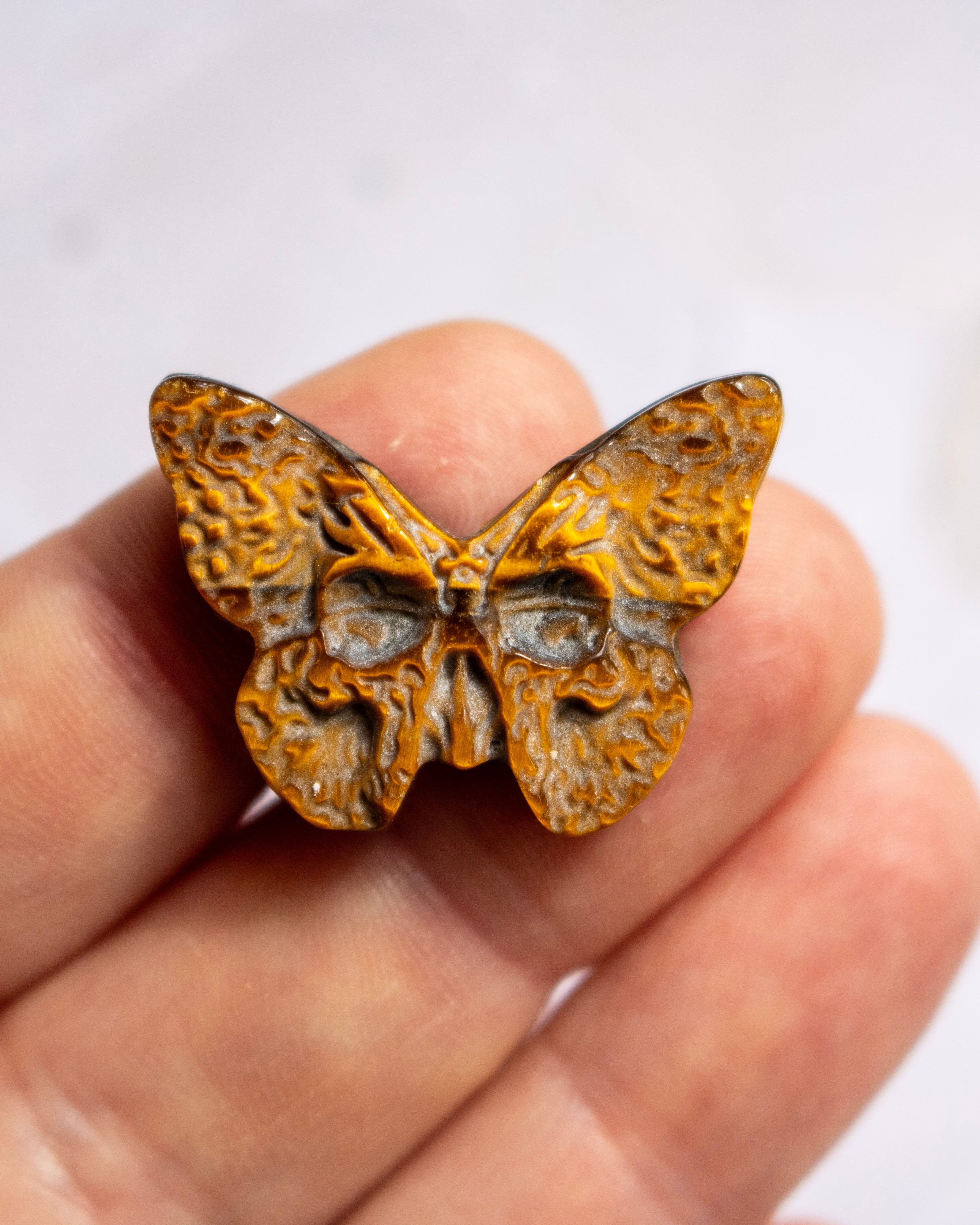 A small carved butterfly-shaped object with textured orange-brown and dark gray details, held between fingers against a soft light background.